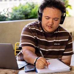 A man with piercings outside on a bench with their laptop and notepad
