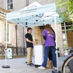 People attending a previous Open Day Two student workers and one visitor at the Open Day standing near the entrance to a University of Cambridge building, under a University of Cambridge branded gazebo