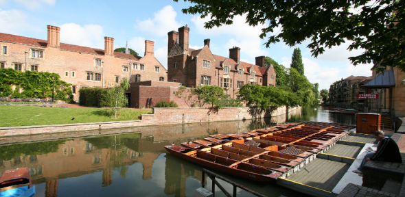 River Court - Magdalene College - University of Cambridge Magdalene College student studying by the river Cam in the shade of a tree, with punts in front of him in the sun