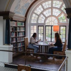 Image of Newnham students at a table in the Newnham Library. One reads while the other is on a laptop. 