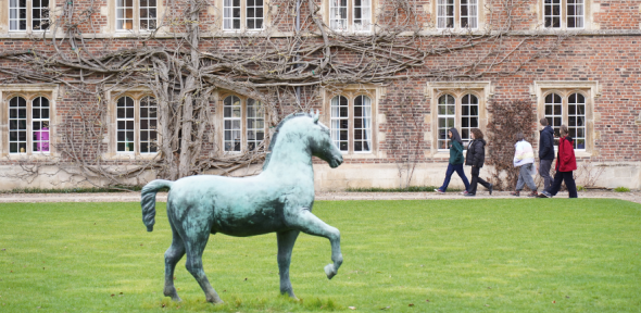A photo of the horse statue at St John's College's garden, and three students walking in the background, facing the windows of the building.